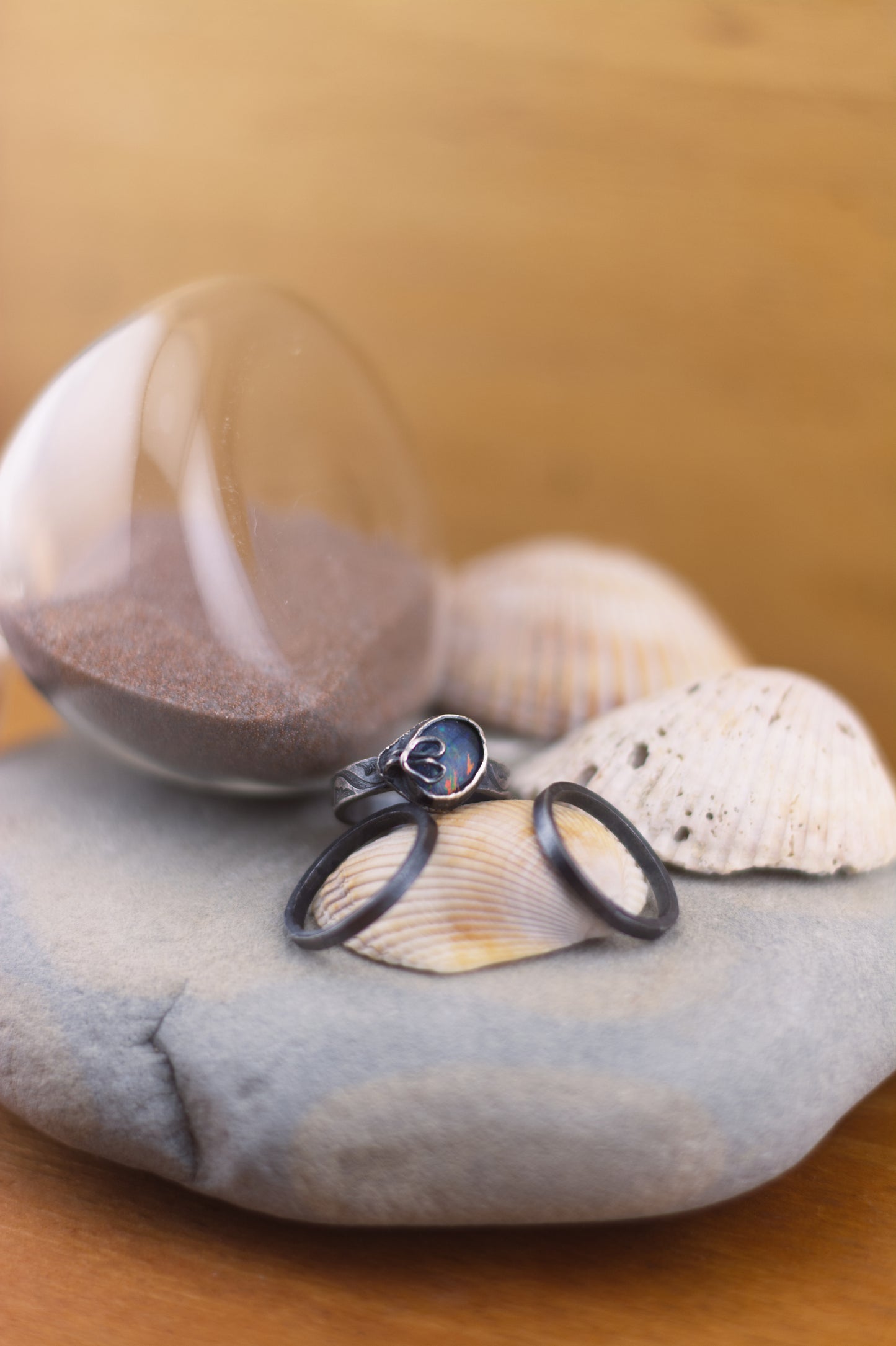 Fairy Rainbow Ring - Three patina'd sterling silver rings. The patterned ring is set with an opal and a beansprout prong. All sitting atop seashells and a rock with half an hour glass on a beige background